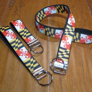 Puede incluir: Tres llaveros con diseños de bandera del estado de Maryland en negro, amarillo y rojo. Los llaveros tienen anillos de llaves plateados.
