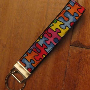 May include: A colorful key chain with a puzzle piece pattern. The key chain is made of fabric and has a black background with red, orange, yellow, green, blue, and purple puzzle pieces. The key chain has a silver metal key ring.