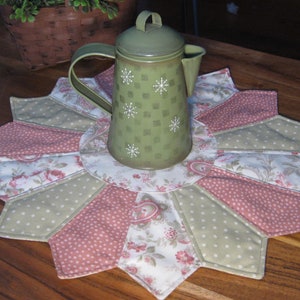 May include: A green metal coffee pot with white snowflake designs sits on a quilted placemat. The placemat features alternating sections of pink polka dots, floral patterns, and light green polka dots. The placemat is on a wooden table.