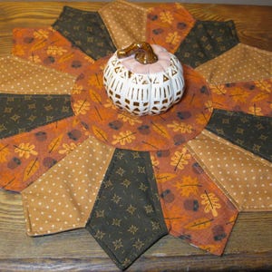 May include: A brown, orange and green fabric table runner with a white ceramic pumpkin in the centre. The runner has a starburst design with a variety of autumn-themed patterns.