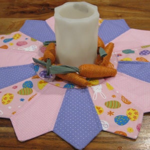 May include: A pink and purple fabric table centerpiece with a white candle in the center. The centerpiece is decorated with carrots and Easter-themed fabric.