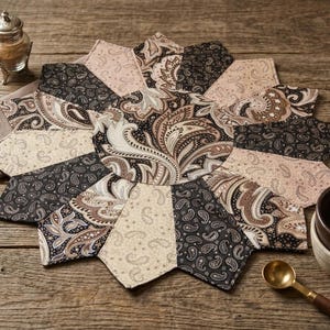 May include: A decorative, star-shaped placemat with a paisley pattern in shades of black, brown, and beige. The placemat is made of fabric and sits on a wooden surface. A small bowl, spoon, and salt shaker are also visible.