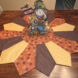 May include: A circular fabric table mat featuring a starburst pattern of alternating brown, yellow, and orange leaf-print panels. Centered on the mat is a decorative metal snowman figurine wearing a hat and holding a sign that reads WELCOME.
