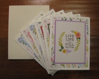 Op de afbeelding: Een set van acht blanco wenskaarten met bloemenpatronen en een paarse rand. De voorkant van de kaart staat "Live life in full bloom".