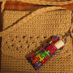 May include: A tan crocheted bag with a long strap. The bag has a zipper closure and a small pocket on the front. A colorful fabric pouch with a zipper is attached to the bag.