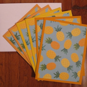 May include: A set of greeting cards with a pineapple pattern. The cards have a light blue background with yellow pineapples and green leaves. The cards have a yellow and orange border and are fanned out next to a white envelope.