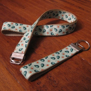 Puede incluir: Un llavero verde y marrón con un patrón de mitades de aguacate. El llavero tiene un cierre de metal plateado y un anillo de metal plateado.