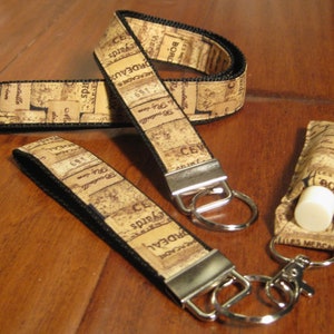 Puede incluir: Una colección de accesorios con estampado de corcho, que incluye dos llaveros con herrajes plateados, un cordón y un soporte para bálsamo labial. Los artículos presentan un ribete negro y texto de etiquetas de vino.