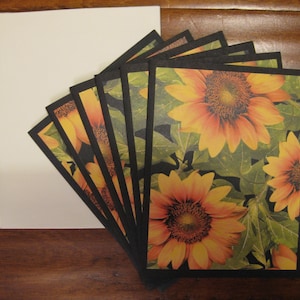 Puede incluir: Un juego de tarjetas de felicitación en blanco con un diseño de girasol. Las tarjetas tienen bordes negros y muestran un primer plano de girasoles con hojas verdes.