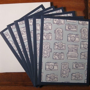 May include: A set of ten blank greeting cards with a blue border and a light blue background. The cards feature a repeating pattern of vintage cameras in black and white.