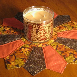 May include: A quilted candle mat with a starburst design in shades of brown, orange, and gold. The mat features a central circle with a decorative candle holder.