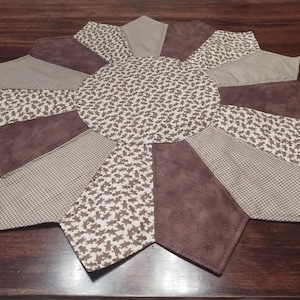 Puede incluir: Un mantel individual artesanal en forma de estrella. El mantel individual presenta un círculo central y segmentos radiales en varios patrones marrones y beige, incluyendo texturas florales, de cuadros y lisas. El mantel individual está diseñado para uso decorativo.