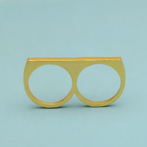 Puede incluir: Un anillo dorado para dos dedos. El anillo tiene dos aberturas circulares y una banda superior rectangular. El anillo está hecho de un material sólido, posiblemente metal, y está sobre un fondo azul claro.