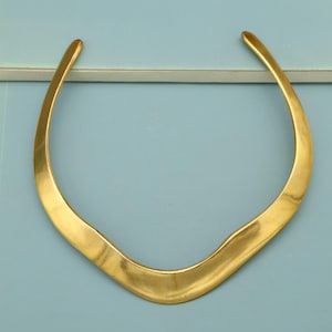 Könnte beinhalten: Eine goldfarbene Halskette mit einem gebogenen, offenen Design. Die Halskette hat eine glatte, polierte Oberfläche und eine leicht unregelmäßige Form. Das Schmuckstück ist vor einem hellblauen Hintergrund platziert.