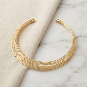 Puede incluir: Un collar tipo gargantilla de tono dorado con un diseño de múltiples hilos compuesto por varias bandas delgadas y paralelas que se curvan en forma circular. El collar tiene extremos cilíndricos lisos en la abertura, creando un aspecto estructurado.