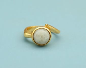 May include: A gold-colored ring with a round, light-colored stone set in the center. The ring has a simple, elegant design with a smooth, polished finish. The stone appears to be a pale, opaque color.