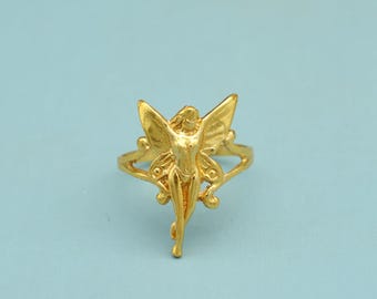 May include: A gold-colored ring featuring a fairy design. The fairy has outstretched wings and is centered on the ring's band. The ring has a detailed, ornate design with a polished finish. The background is a solid light blue.