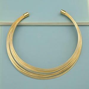 May include: A gold-colored necklace with multiple strands. The necklace has a choker style and is made of thin, flexible wires. The ends of the necklace have a gold-colored clasp. The background is a light blue color.