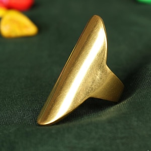 Op de afbeelding: Een goudkleurige ring met een lang, puntig ontwerp. De ring heeft een glad, gepolijst oppervlak en een band die om de vinger gaat. De ring staat tegen een donkergroene achtergrond.