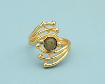 Op de afbeelding: Een goudkleurige ring met een centrale, ronde, olijfgroene steen. De ring heeft een open ontwerp met meerdere gebogen banden en kleine, bolvormige accenten. De ring is geplaatst tegen een lichtblauwe achtergrond.