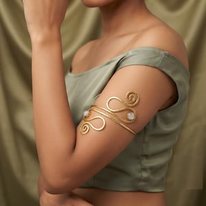 May include: A gold-toned metal arm cuff featuring a textured, swirling wire design that wraps around the upper arm. The piece is accented with two small, round, iridescent white cabochon stones set within the metal spirals on the front.