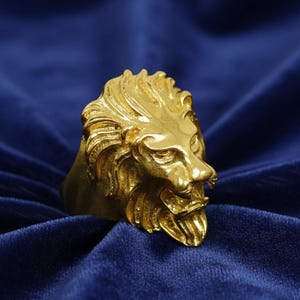 May include: A gold-colored ring featuring a detailed lion's head design. The lion's mane and facial features are intricately sculpted, showcasing a regal and powerful aesthetic. The ring is set against a deep blue background.