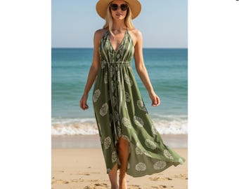 Könnte beinhalten: Eine Frau trägt ein olivgrünes Maxikleid mit Neckholder, weißem Blumenmuster, Strohhut und Sonnenbrille an einem Sandstrand. Das Kleid hat einen High-Low-Saum und eine geraffte Taille. Der Hintergrund zeigt das Meer und einen klaren blauen Himmel.