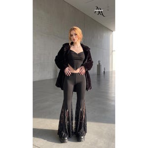 May include: A black outfit consisting of a cropped, pointed-hem top and high-waisted flared pants. The pants feature intricate black lace inserts at the lower legs, accented with small silver cross charms. A black faux fur jacket is worn over the top.