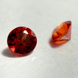 May include: Two round-cut gemstones with multiple facets. One is viewed from above, showing a deep red colour. The other gem is viewed from the side, displaying orange and red hues. The gems are set against a white background.