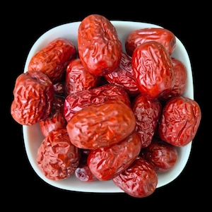 Könnte beinhalten: Eine weiße Schale gefüllt mit einer Ansammlung getrockneter roter Jujube-Früchte. Jede Frucht hat eine faltige, tiefrote Schale und eine ovale Form. Die Jujuben sind in der Schale gestapelt und zeigen ihre vielfältigen Texturen und Farben.