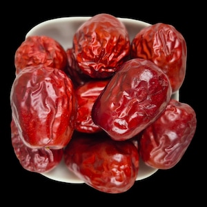 Puede incluir: Un cuenco blanco lleno de varias dátiles rojos sobre un fondo negro. Los dátiles tienen una textura arrugada y un brillo brillante. La imagen es un primer plano, que destaca el color y la textura de la fruta.