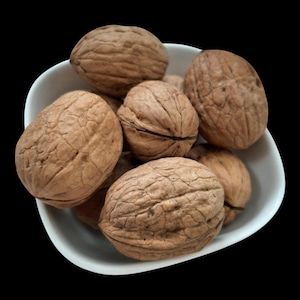 Puede incluir: Un cuenco blanco lleno de varias nueces marrones. Las nueces tienen cáscaras texturizadas y estriadas y están dispuestas en primer plano sobre un fondo negro. La imagen resalta el color y la textura naturales de las nueces.