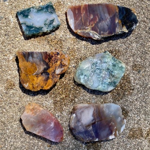May include: Six agate slices with various patterns and colors, including brown, white, green, and purple. The agate slices are arranged on a light brown, sandy surface.
