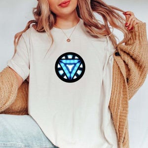 May include: A cream-colored t-shirt featuring a circular graphic with a blue and white triangle design. The graphic is centered on the front of the shirt. The shirt has short sleeves and a crew neck.