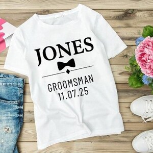 Może przedstawiać: Biała koszulka z napisem "JONES" i słowem "GROOMSMAN" wydrukowanym na czarno, wraz z motywem muchy i datą "11.07.25". Koszulka jest prezentowana z niebieskimi dżinsami, pudełkiem prezentowym z różową kokardą i białymi trampkami.