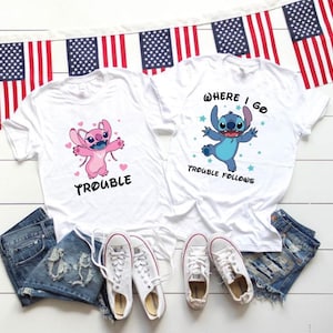 Peut inclure: Deux t-shirts blancs à manches courtes. Le t-shirt de gauche présente un personnage rose avec des cœurs et le texte "TROUBLE". Celui de droite présente un personnage bleu avec des étoiles et le texte "WHERE I GO TROUBLE FOLLOWS". Ils sont accompagnés de shorts en jean et de baskets blanches.