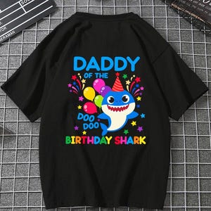 Op de afbeelding: Een zwart t-shirt met korte mouwen en een kleurrijke afbeelding van een blauwe haai met een feesthoedje op. De tekst luidt DADDY OF THE BIRTHDAY SHARK en DOO DOO, omringd door illustraties van veelkleurige ballonnen, sterren en vuurwerk.