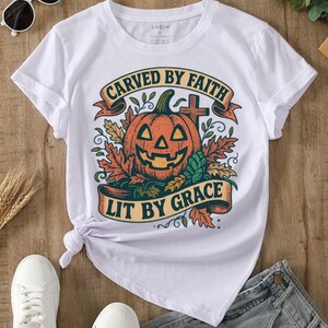 Op de afbeelding: Wit T-shirt met een Halloween-ontwerp. De afbeelding toont een oranje pompoen, herfstbladeren en de tekst "CARVED BY FAITH LIT BY GRACE" op een banner. Het T-shirt wordt gecombineerd met een spijkerbroek en witte sneakers.