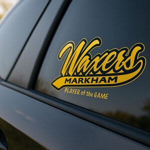 May include: A yellow and black sticker on a car window. The sticker features the word "Waxers" in a stylized font, with "MARKHAM" and "PLAYER of the GAME" below it. The car is dark gray.