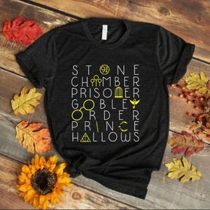 Peut inclure: T-shirt gris foncé avec texte blanc et jaune et des symboles. Le t-shirt est entouré de feuilles d'automne et d'un tournesol sur une surface en bois. Le texte comprend les mots "Stone", "Chamber", "Prisoner", "Goblet", "Order", "Prince" et "Hallows".