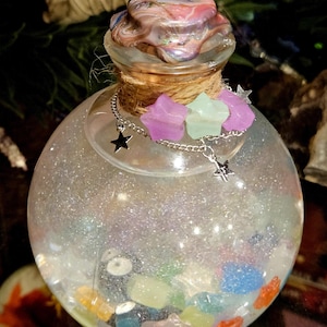 Peut inclure: Une bouteille en verre ronde et transparente remplie de paillettes et de pierres en forme d'étoiles colorées. La bouteille a un couvercle décoratif et est ornée d'une chaîne argentée et de breloques en forme d'étoiles.