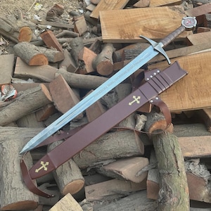 May include: A long, silver sword with a brown handle and a cross-shaped guard, resting in a brown leather sheath. The sheath has a gold cross emblem. The sword's blade has etched text. The sword is approximately 40 inches long.