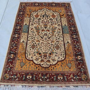 Pode incluir: Tapete retangular com um campo central creme apresentando um design floral ornamentado em tons de laranja, verde e castanho. O tapete tem uma borda vermelha escura com padrões repetidos e franjas. Provavelmente feito de lã.