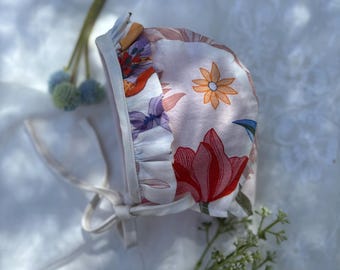 May include: A floral print baby bonnet with a ruffled edge and tie closure. The bonnet features a pattern of red tulips, orange and purple flowers on a light pink background. The ties are a cream color.
