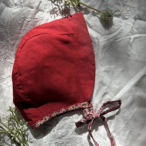 May include: A red bonnet with a floral patterned trim and matching ties. The bonnet is made of a textured fabric and is likely handmade. The ties are tied in a bow. The bonnet is laying on a white, textured surface.