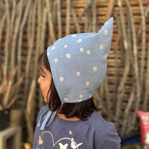 May include: A light blue bonnet with a white daisy pattern. The bonnet has a pointed top and ties under the chin. The child is wearing a blue long-sleeved shirt with a heart and butterfly design.