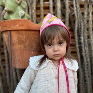 May include: A child wearing a pink bonnet with a yellow lemon pattern and a cream-colored dress with a white collar. The dress has small floral details and wooden buttons. A pink ribbon ties the bonnet.