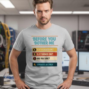 Puede incluir: Camiseta gris claro con el texto "BEFORE YOU BOTHER ME" en azul y naranja. Debajo, cuatro pasos de solución de problemas numerados en cajas de colores. La camiseta la lleva una persona.