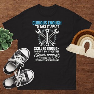 以下が含まれることがあります： 「CURIOUS ENOUGH TO TAKE IT APART, SKILLED ENOUGH TO PUT IT BACK TOGETHER, Clever enough TO HIDE THE EXTRA PARTS WHEN I'M DONE」のテキストが入った黒いTシャツ。デザインには交差したレンチが含まれています。