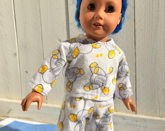 May include: A doll in a white pajama set patterned with grey elephants and yellow details. The doll has blue hair. The pyjama set includes a top and trousers. The doll is standing on a blue surface.